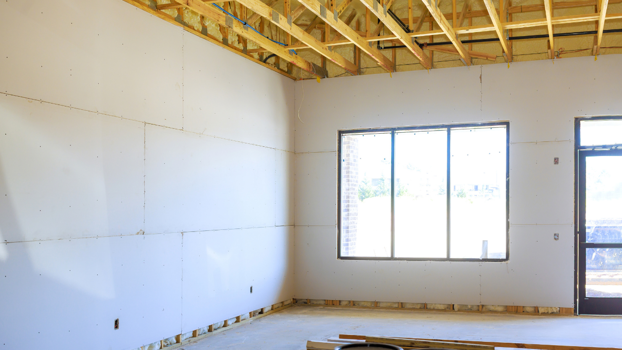 Interior of a building under construction, drywall on walls, exposed wood ceiling, window with multiple panes.