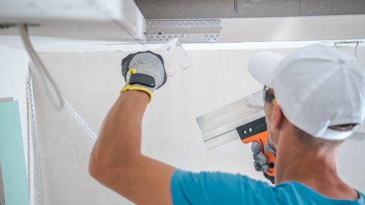 Person wearing gloves and a cap applying drywall compound with a trowel.