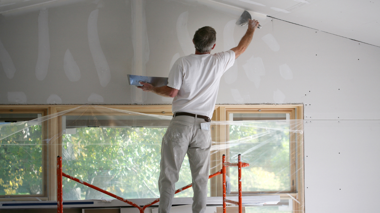 Man on a scaffold applying drywall mud to a wall inside a building.