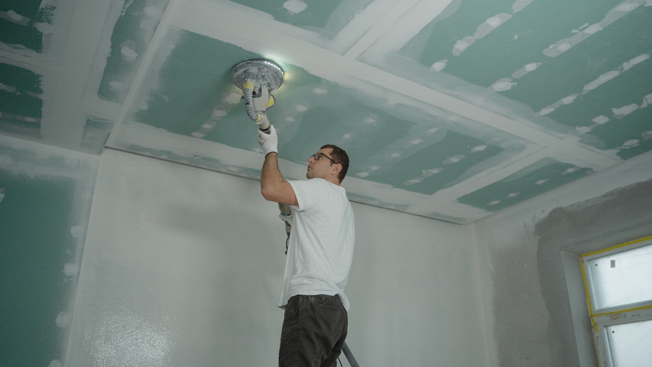Man sanding a freshly plastered ceiling with a handheld sanding machine. White paint and green drywall in a room.