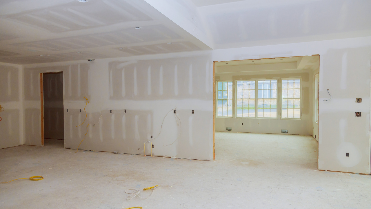 Interior of a room under construction, with drywall walls and ceiling, and a doorway to another room with windows.