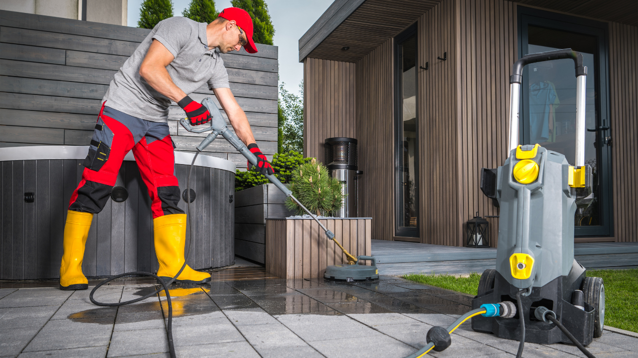 Man pressure washing a wooden deck outdoors, wearing red and yellow work clothes.
