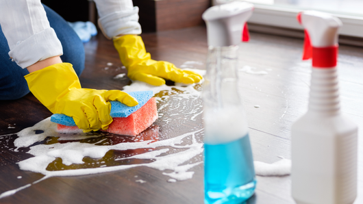 Person wearing yellow gloves, cleaning wooden floor with a sponge and spray bottles.