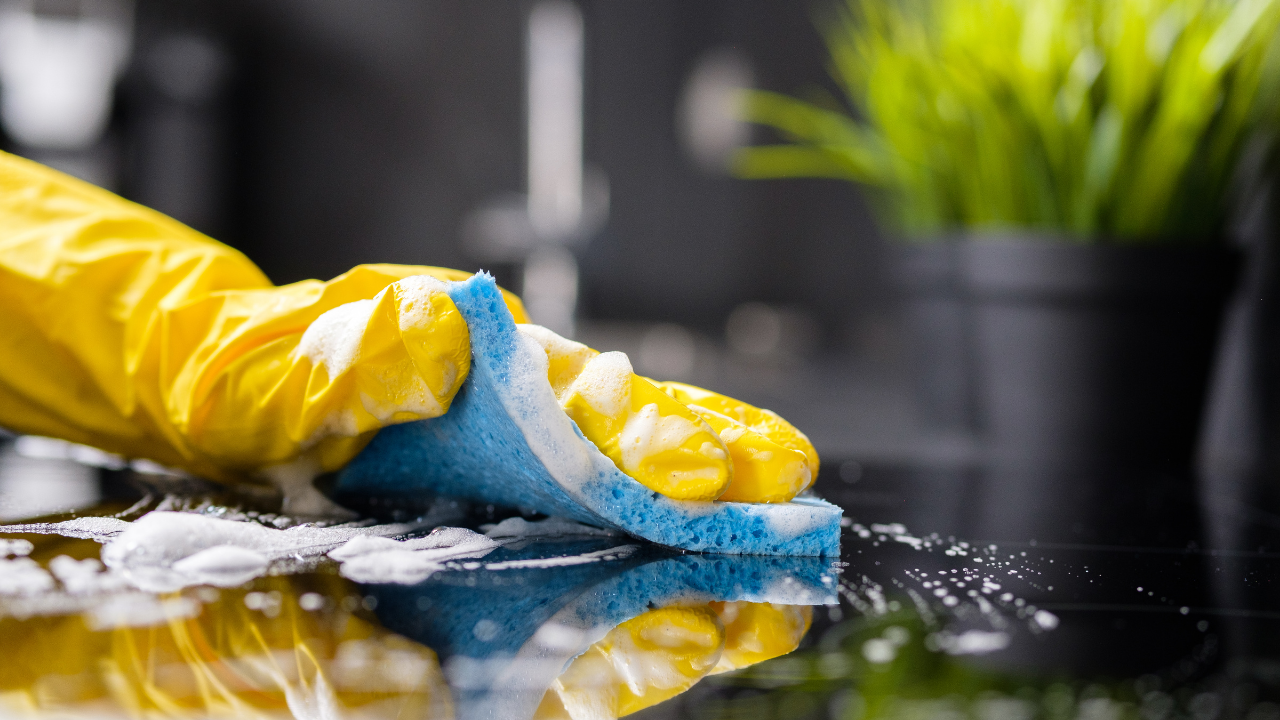 Yellow-gloved hand wiping a dark surface with a blue and white sponge, cleaning the kitchen.