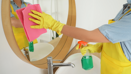Person wearing yellow gloves cleans a bathroom mirror with a pink cloth and spray bottle.