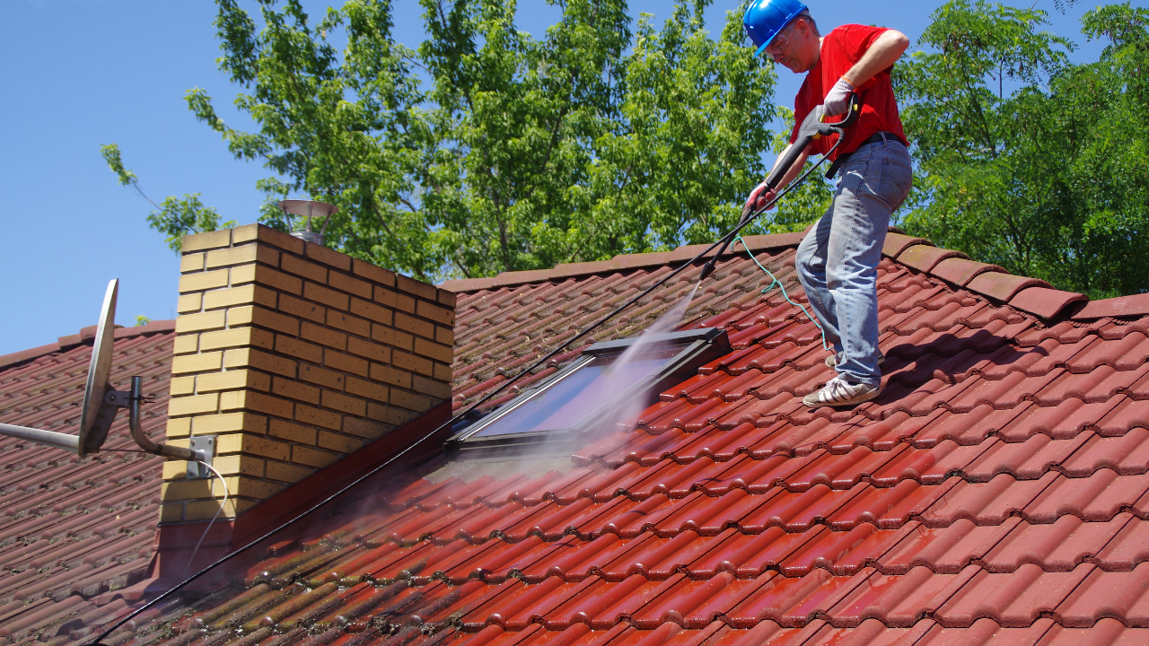 Man in red shirt and blue hard hat pressure-washing a red tile roof, near a brick chimney and skylight.