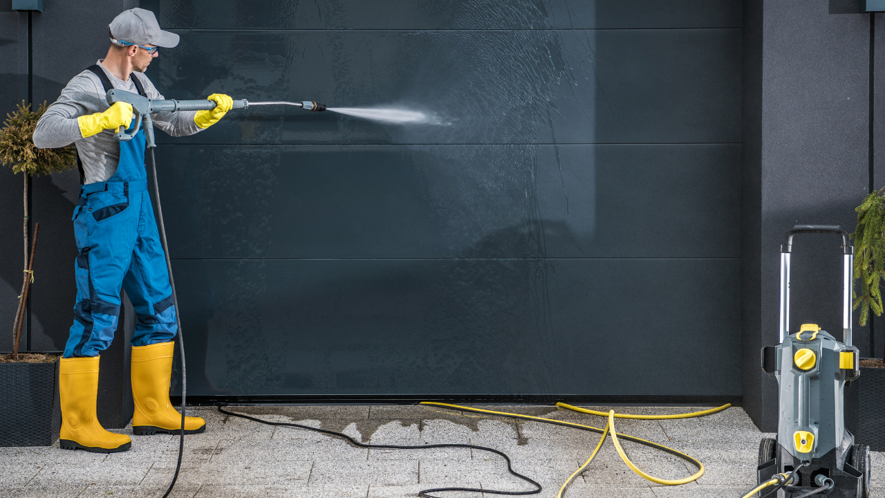 Man pressure washing a dark garage door; yellow boots and gloves, grey water spray.