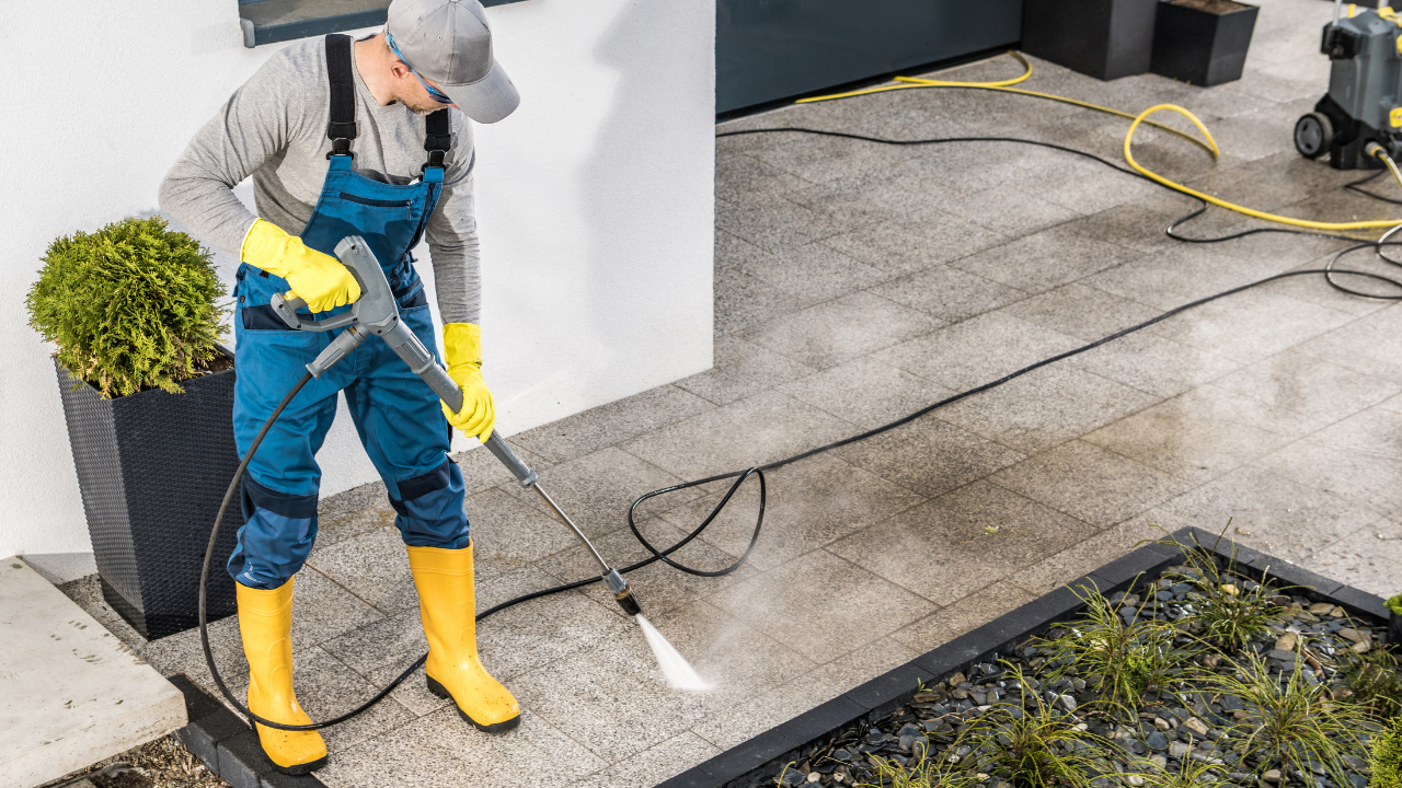 Person power washing a stone patio, wearing overalls, gloves, and boots.