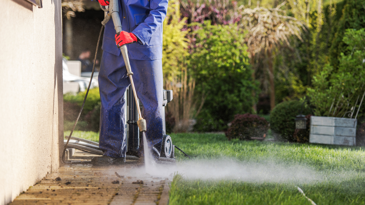 Person in blue overalls power washing a brick patio next to a lawn.