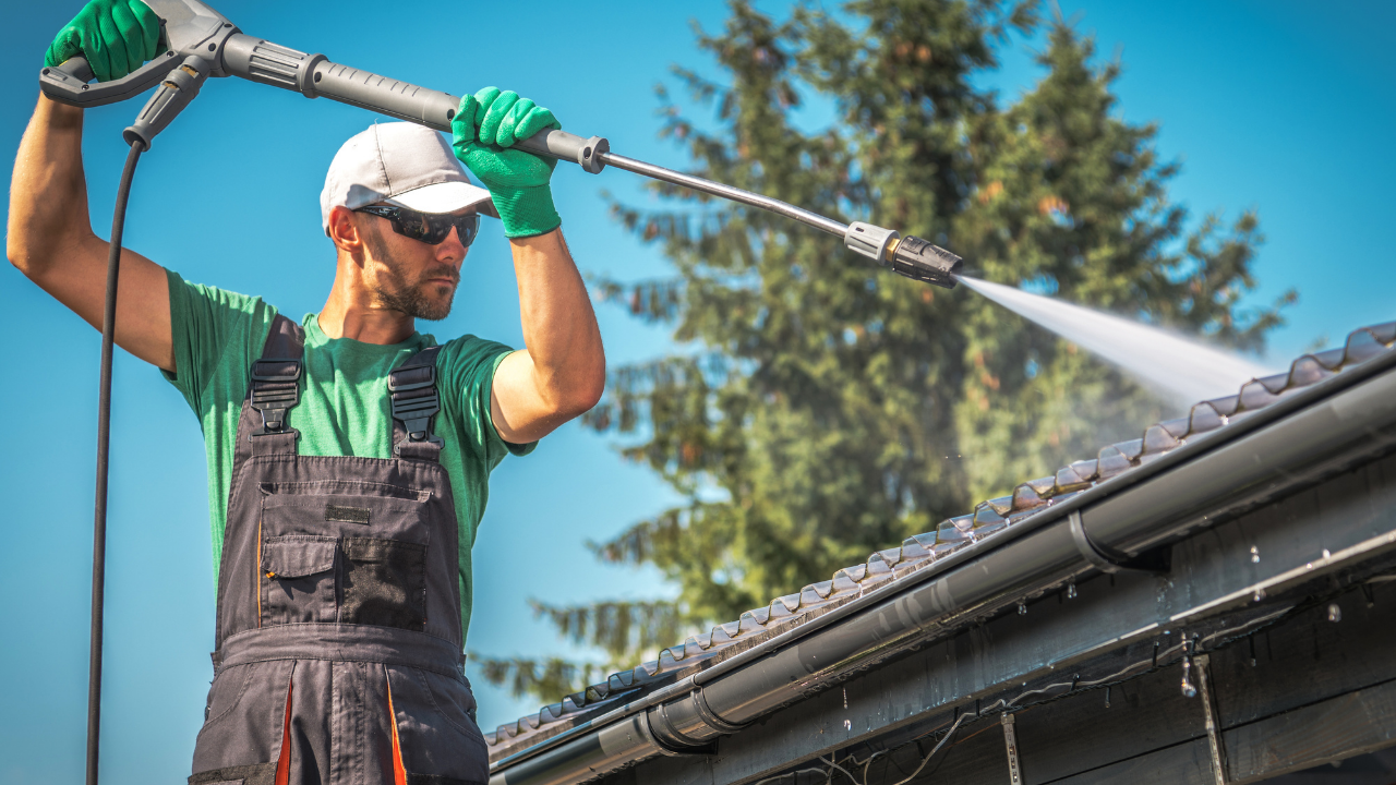 Man in work clothes power washing a dark roof. Green gloves, sunglasses, and a white cap are visible.