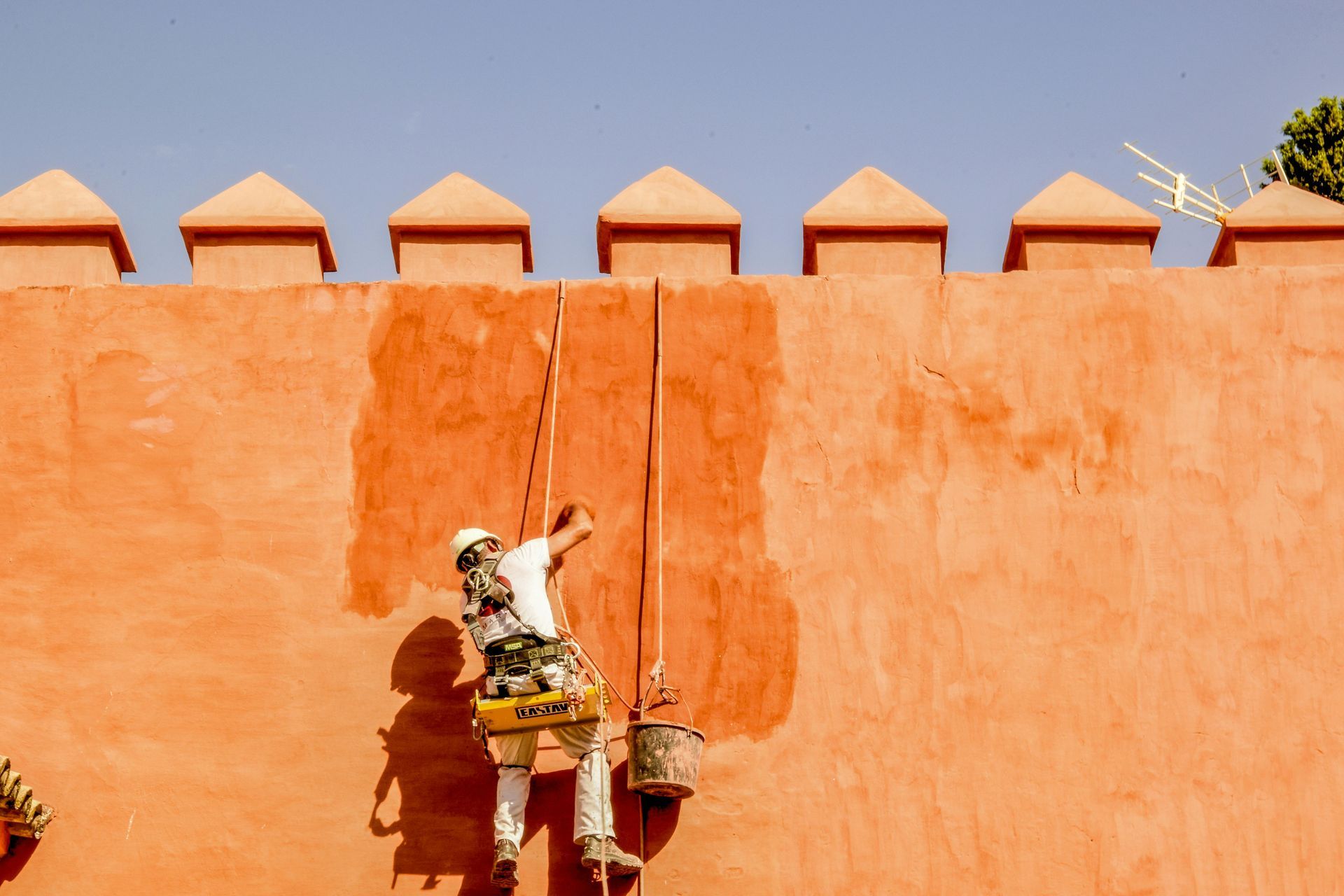 Painter on a rope fixing a crack in an orange wall with a crenelated top, against a blue sky.