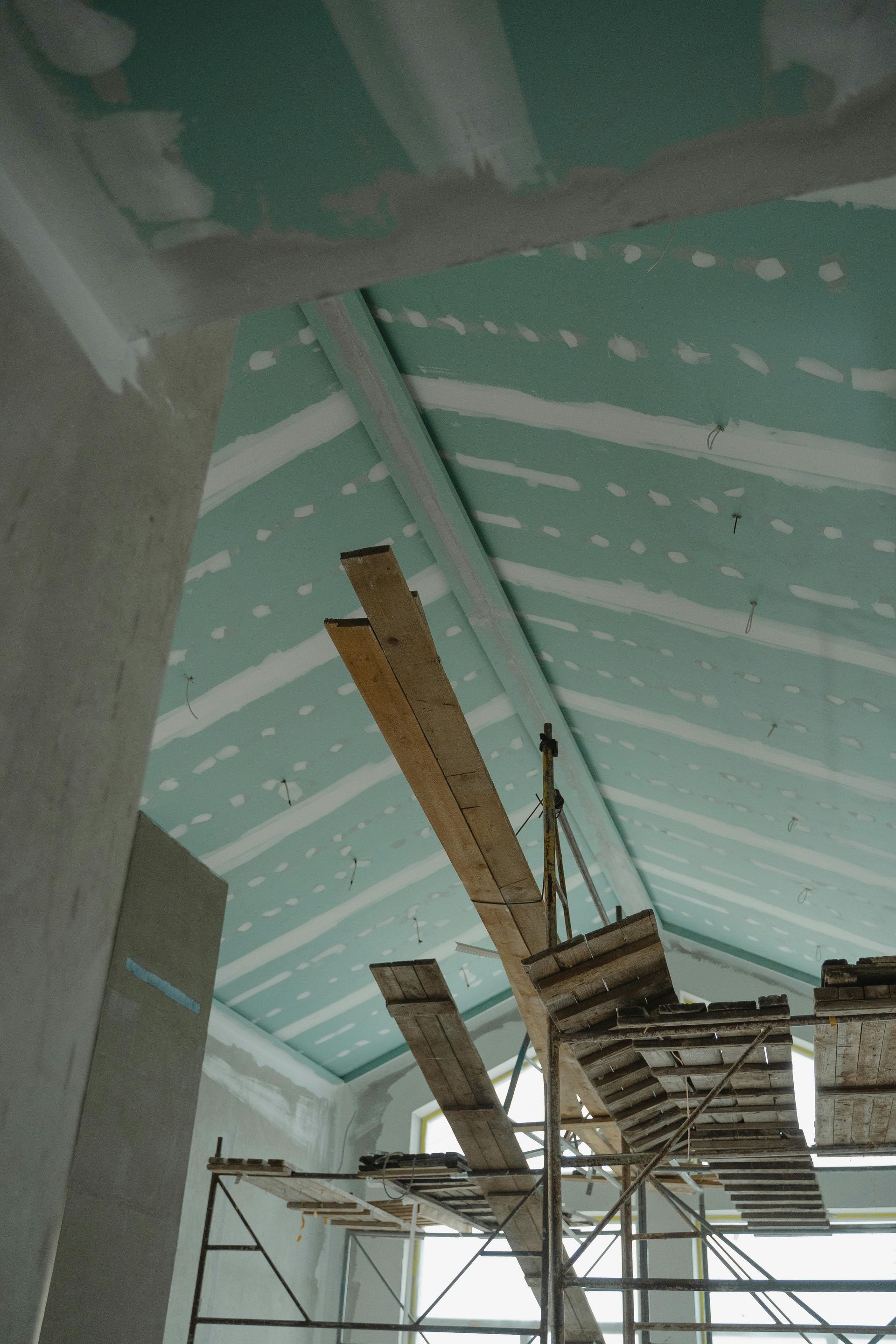 Interior of a building under construction. Green drywall on angled ceiling, scaffolding with wooden planks below.