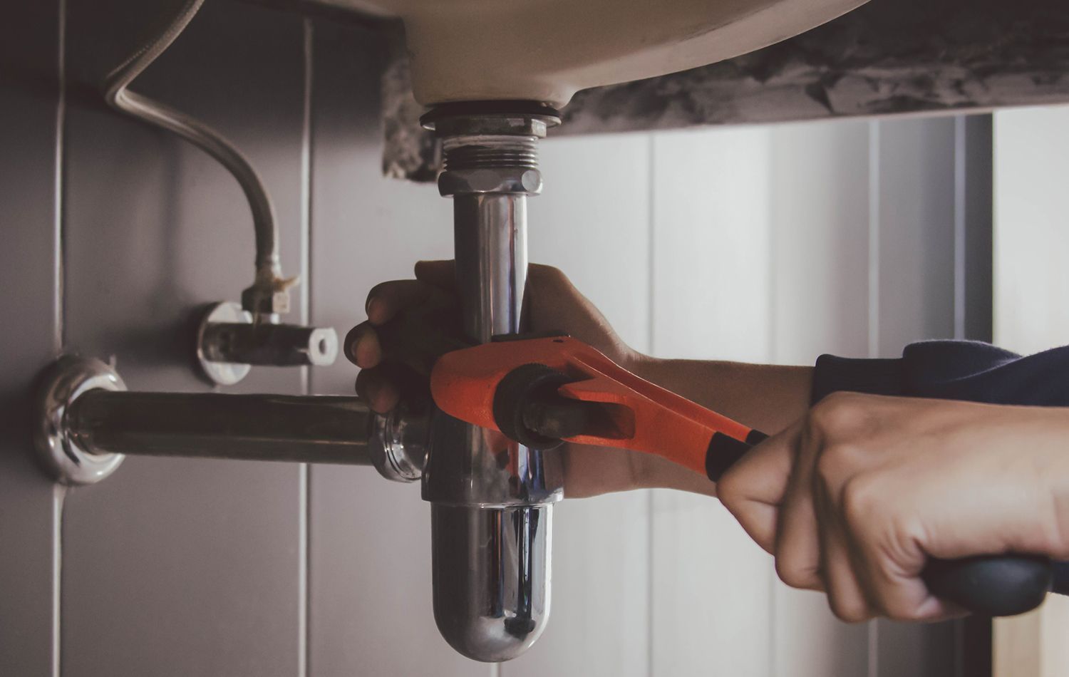 A plumber in work clothes kneeling in a bathroom, using a wrench to tighten the pipe underneath a wall-mounted sink.