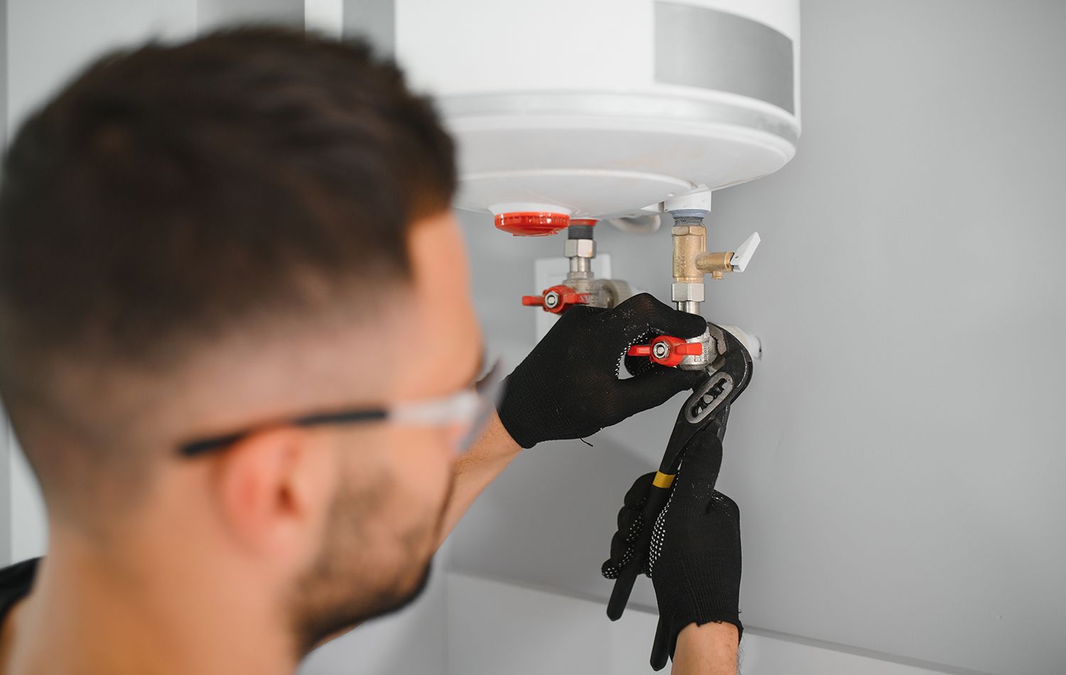 A plumber in work clothes kneeling in a bathroom, using a wrench to tighten the pipe underneath a wall-mounted sink.