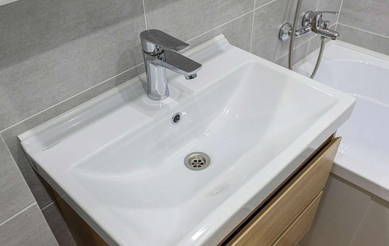 A plumber in work clothes kneeling in a bathroom, using a wrench to tighten the pipe underneath a wall-mounted sink.