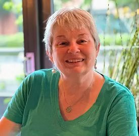Smiling woman in a teal shirt sitting indoors by a window with greenery outside