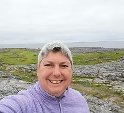 Smiling person in a purple jacket taking a selfie by a rocky coastline under an overcast sky