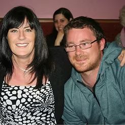 Two smiling people posing indoors, with another person blurred in the background.