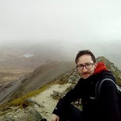 Person in glasses and black hoodie sits on a rocky mountain ridge overlooking a foggy valley.