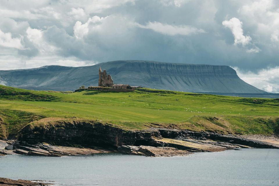 Green coastal cliffs with a small stone ruin and a flat-topped mountain under cloudy skies