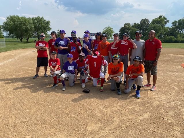 Baseball team posing on a dirt field. Players wear various colored jerseys; some kneel. Trees and blue sky in background.
