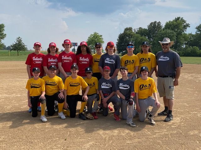 Baseball team photo on a dirt field; boys in red/yellow/gray uniforms, hats, and a coach in a gray shirt, smiling.