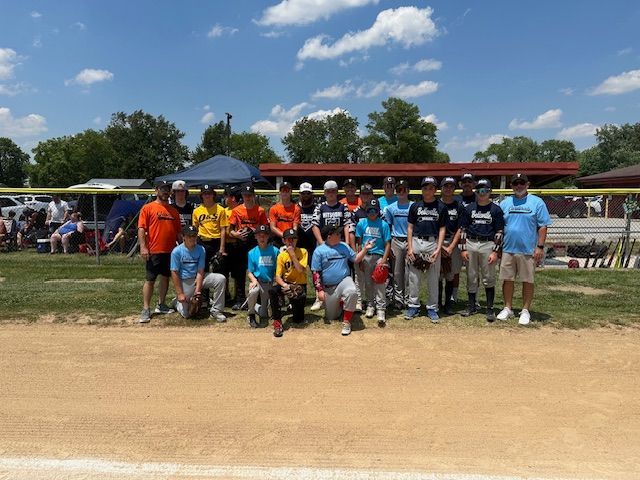 Baseball team posing on a field, some kneeling. Boys wearing jerseys in blue, orange, and black. Sunny day.