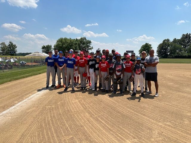 Baseball teams pose together on the field; players in red and blue uniforms stand on the dirt, with coaches.