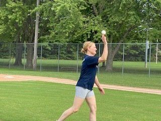 Woman throwing a baseball in a park, wearing shorts and a dark shirt.