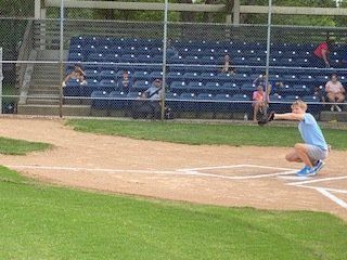 A baseball player squats, catching the ball near home plate, bleachers in the background.