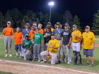Baseball team posing on the field at night. Players in various colored uniforms, some kneeling, under stadium lights.
