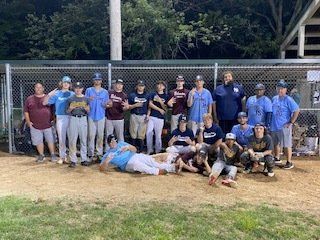 Baseball team poses on field. Players in various uniforms, some kneeling/sitting. Outdoor setting, green trees.