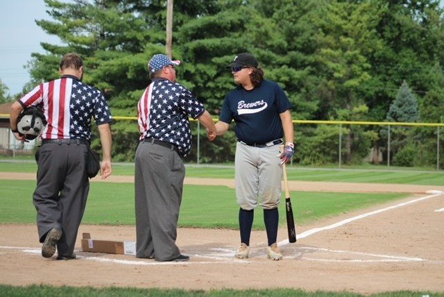 Two umpires in patriotic shirts shake hands with a baseball player wearing Braves gear on a field.