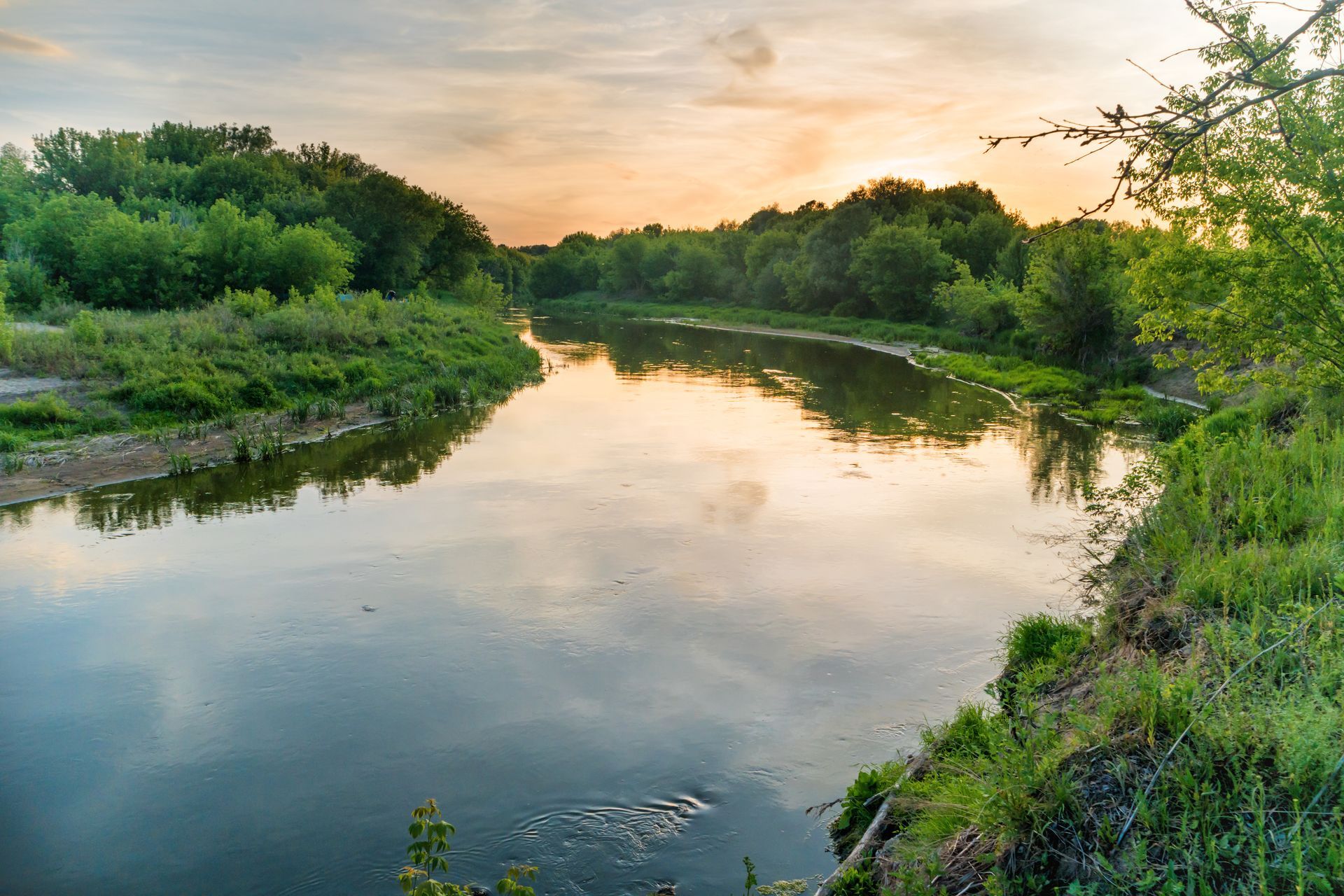 A calm river flows through a lush green landscape at sunset, with light reflecting off the water's surface.
