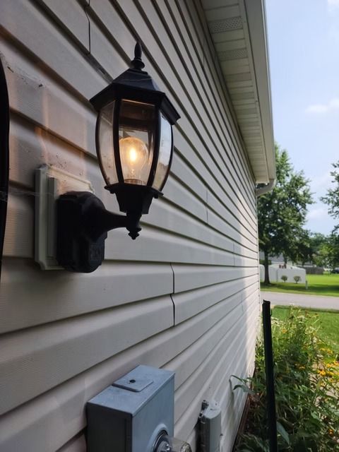 Black outdoor lamp fixture mounted on a cream-colored, horizontal siding wall. A bright light bulb illuminates the lamp.