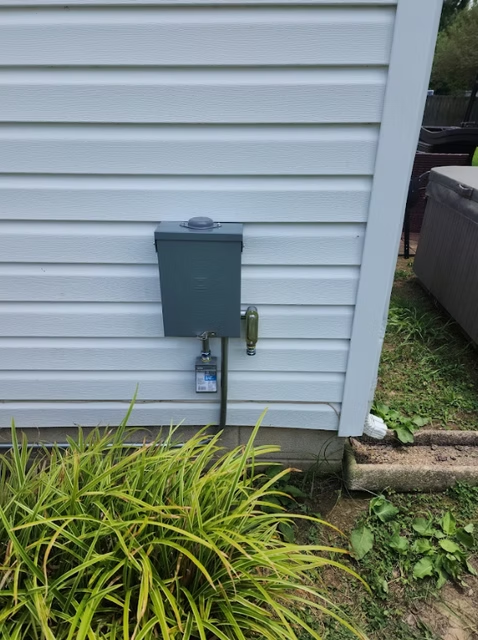 A green electrical box on a light blue-sided building, with a water spigot and a small, clear plastic outlet box.