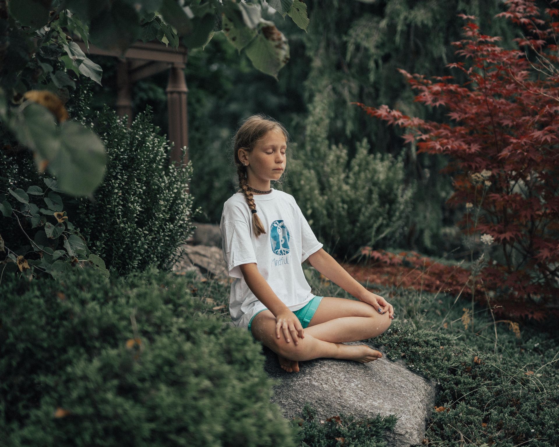 A young girl with braided hair meditating on a rock, surrounded by trees and shrubs