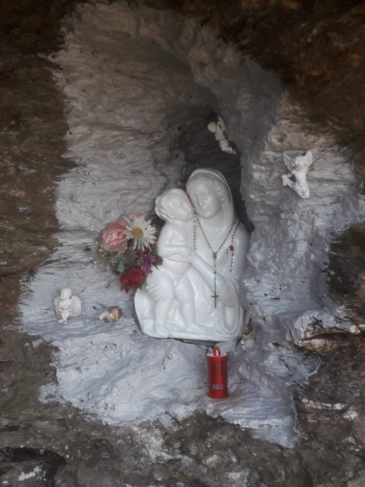 Una statua bianca di Maria con il bambino in una nicchia di pietra con fiori e una candela.