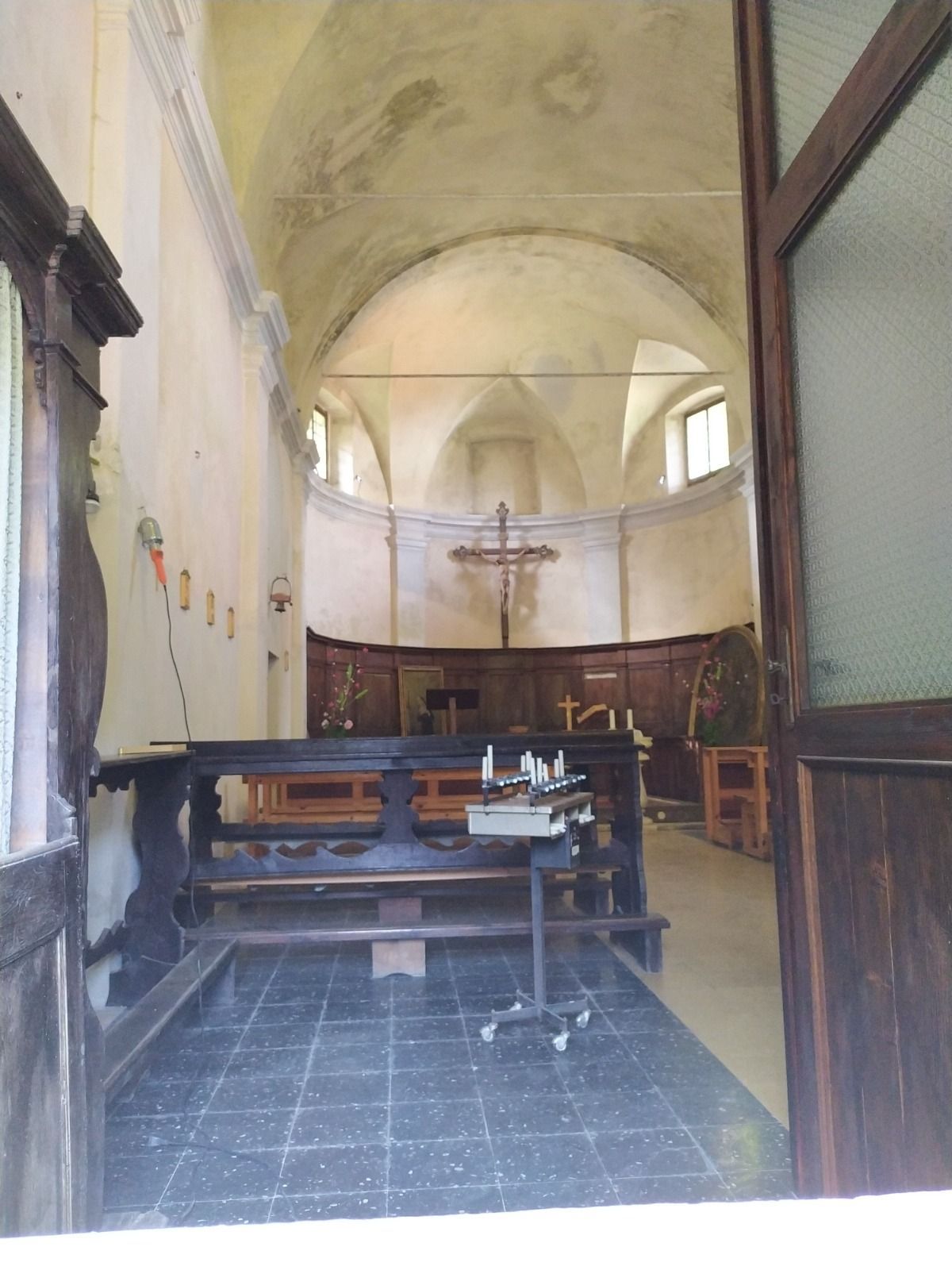 Interno di una chiesa visto attraverso una porta aperta. Soffitto a volta, altare con, panche in legno scuro e pavimento.