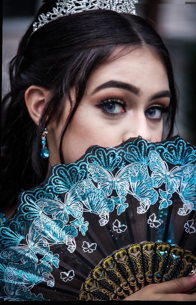 Woman holding a blue-and-black patterned fan over her mouth, with dramatic eye makeup and a dark background
