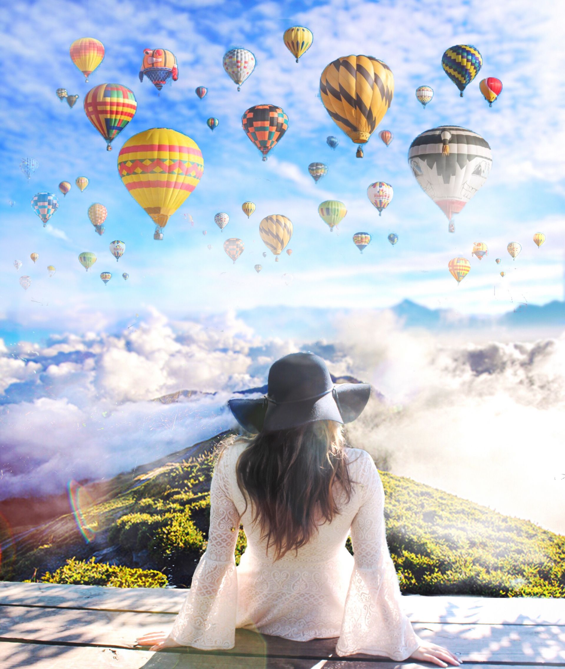 Person seated on a cliff overlooking clouds and colorful hot air balloons in a bright sky