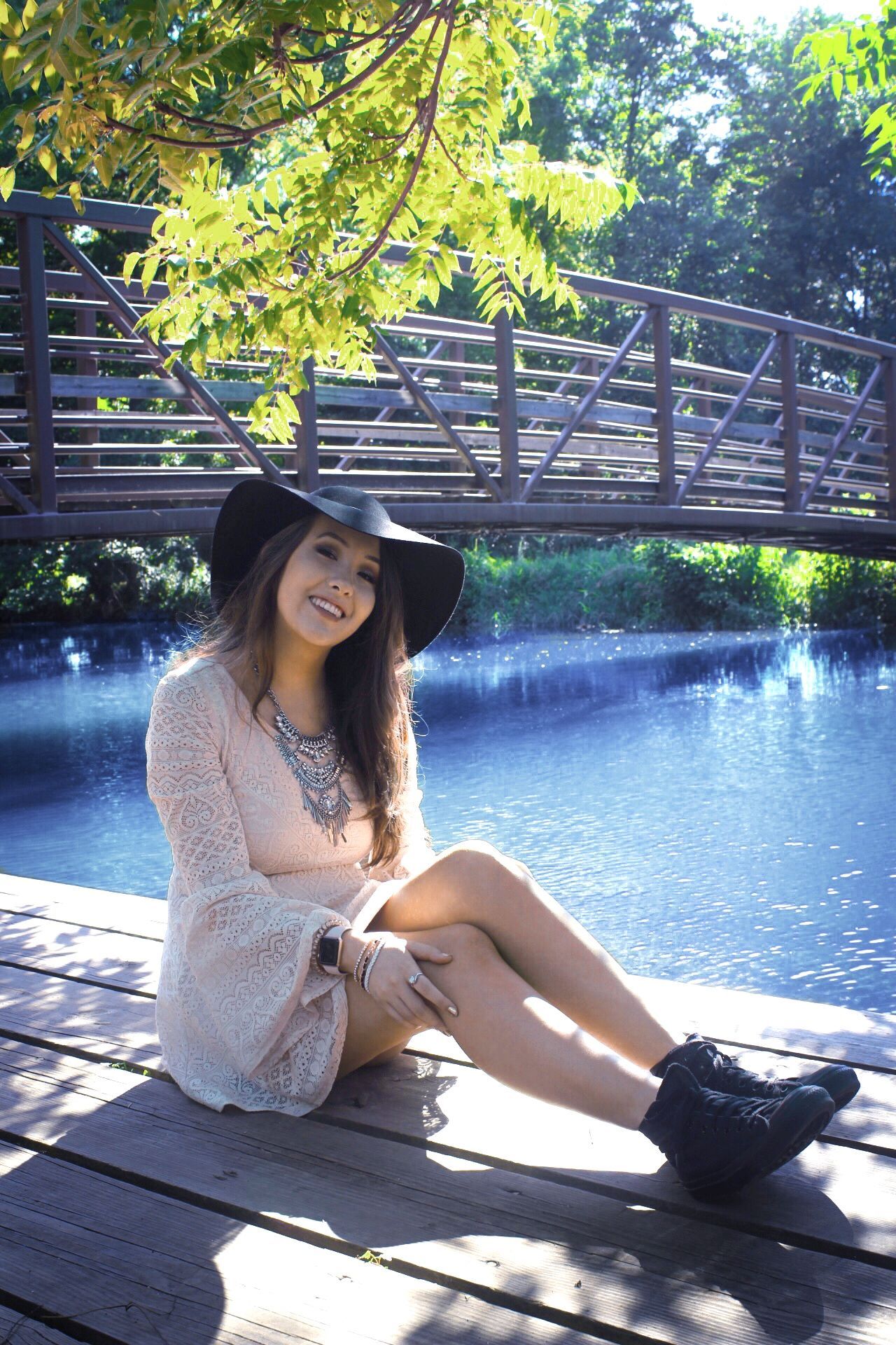 Woman in a black hat sitting on a wooden deck by a blue lake, with a bridge and trees behind her.