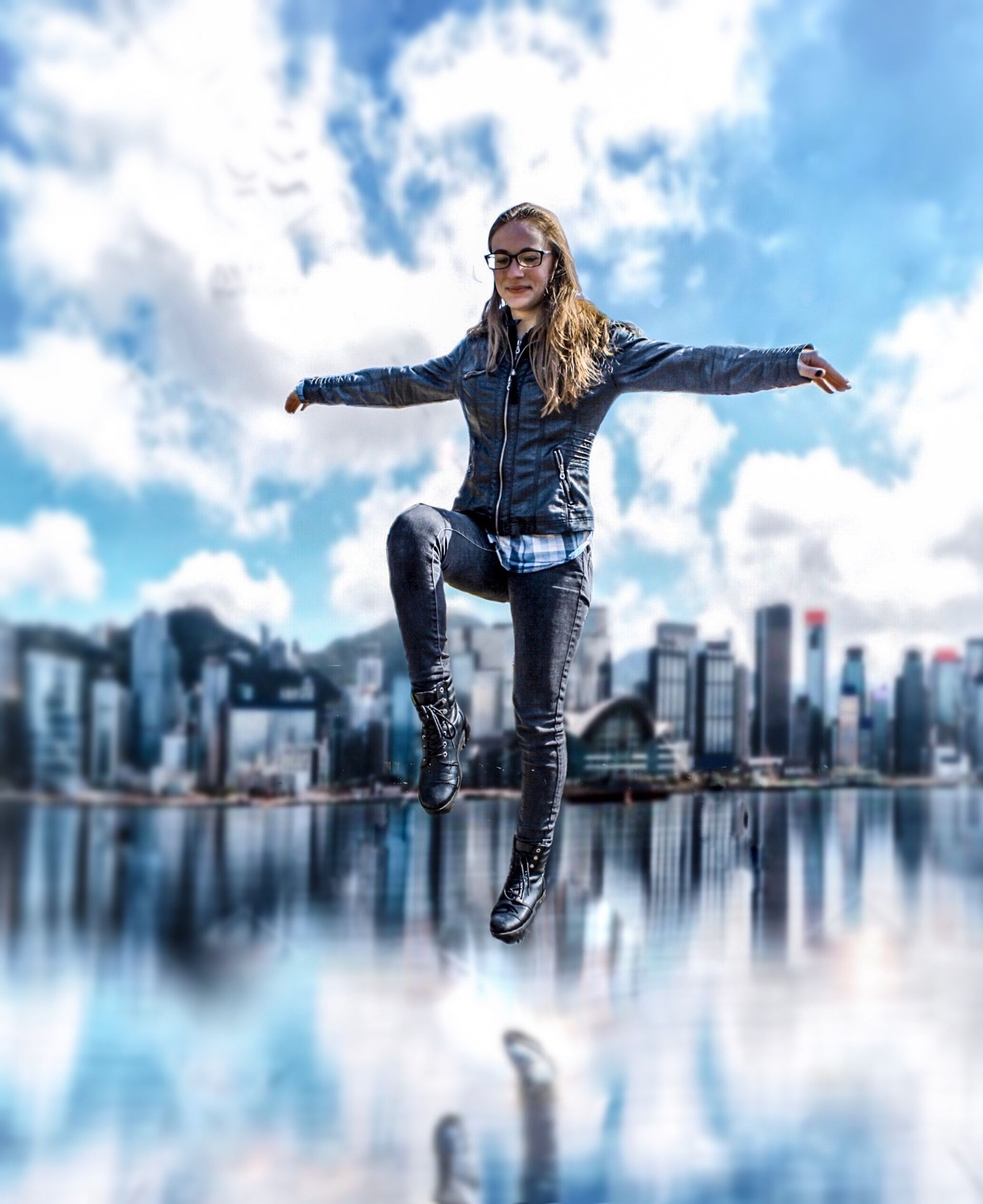 Person in black clothing balancing on one leg before a city skyline reflected in water under a cloudy blue sky