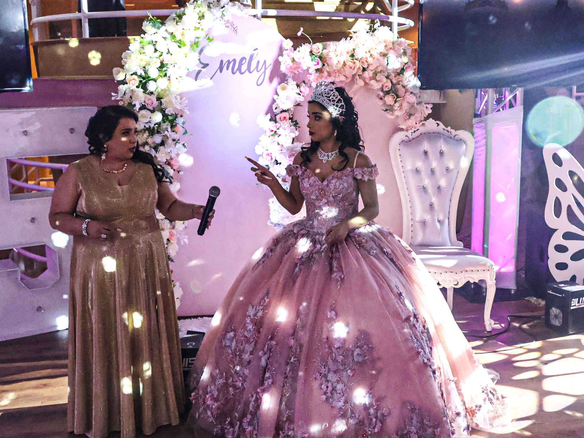 Two women in sparkling gowns pose at a decorated event backdrop with pink flowers and lights