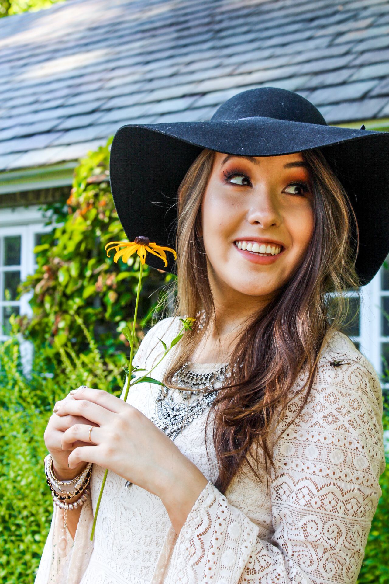 Woman in a black hat smiling while holding a yellow flower outdoors by a house