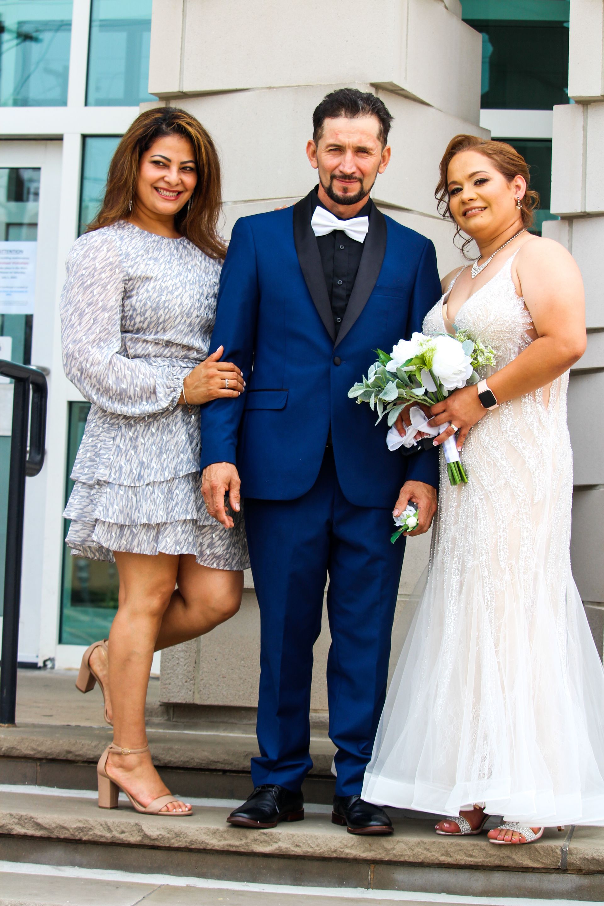 Three people posing outside, with one in a blue suit and one in a white dress holding flowers.