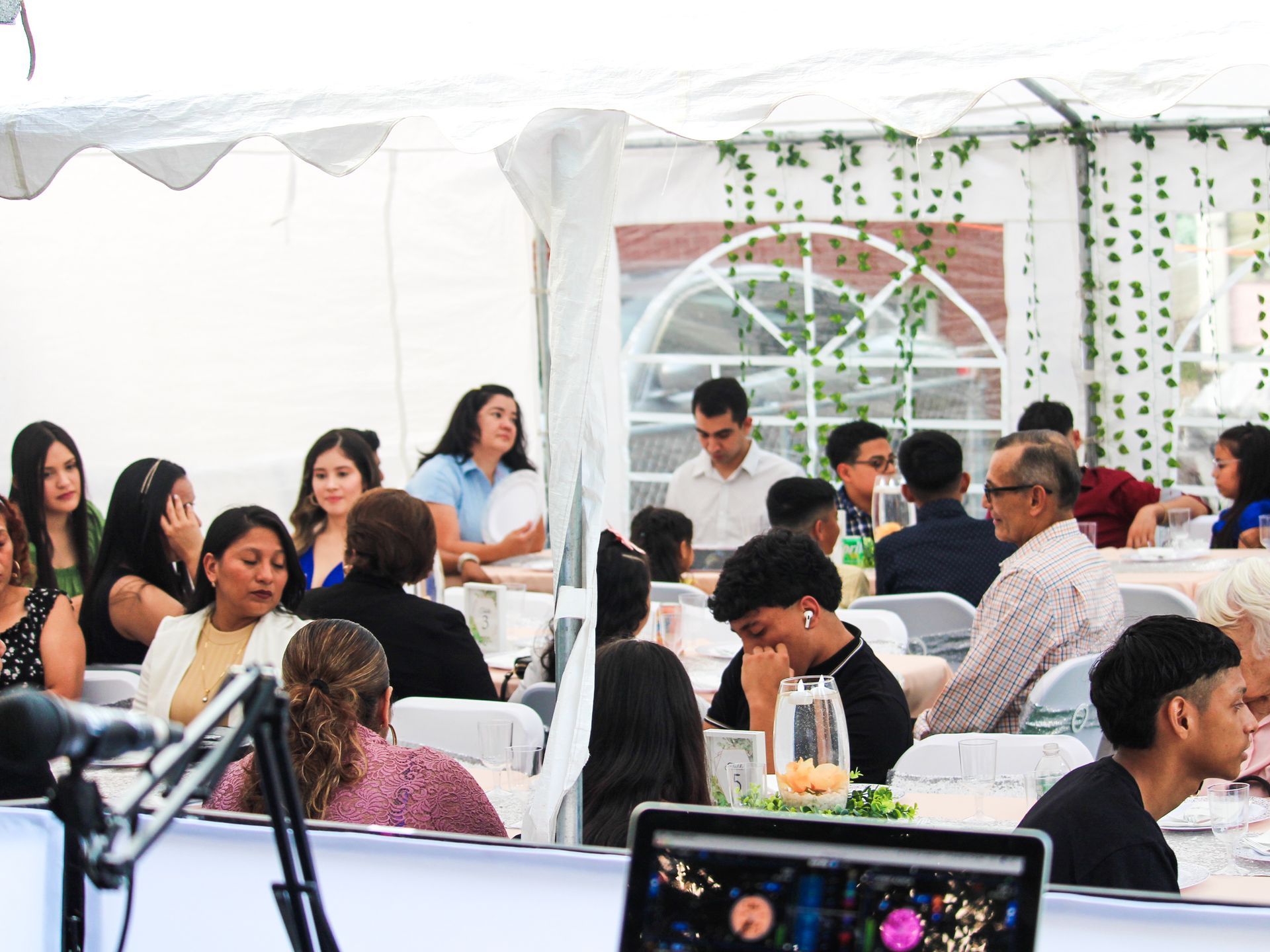 People seated at tables under a white tent at an outdoor event, with a laptop in the foreground.