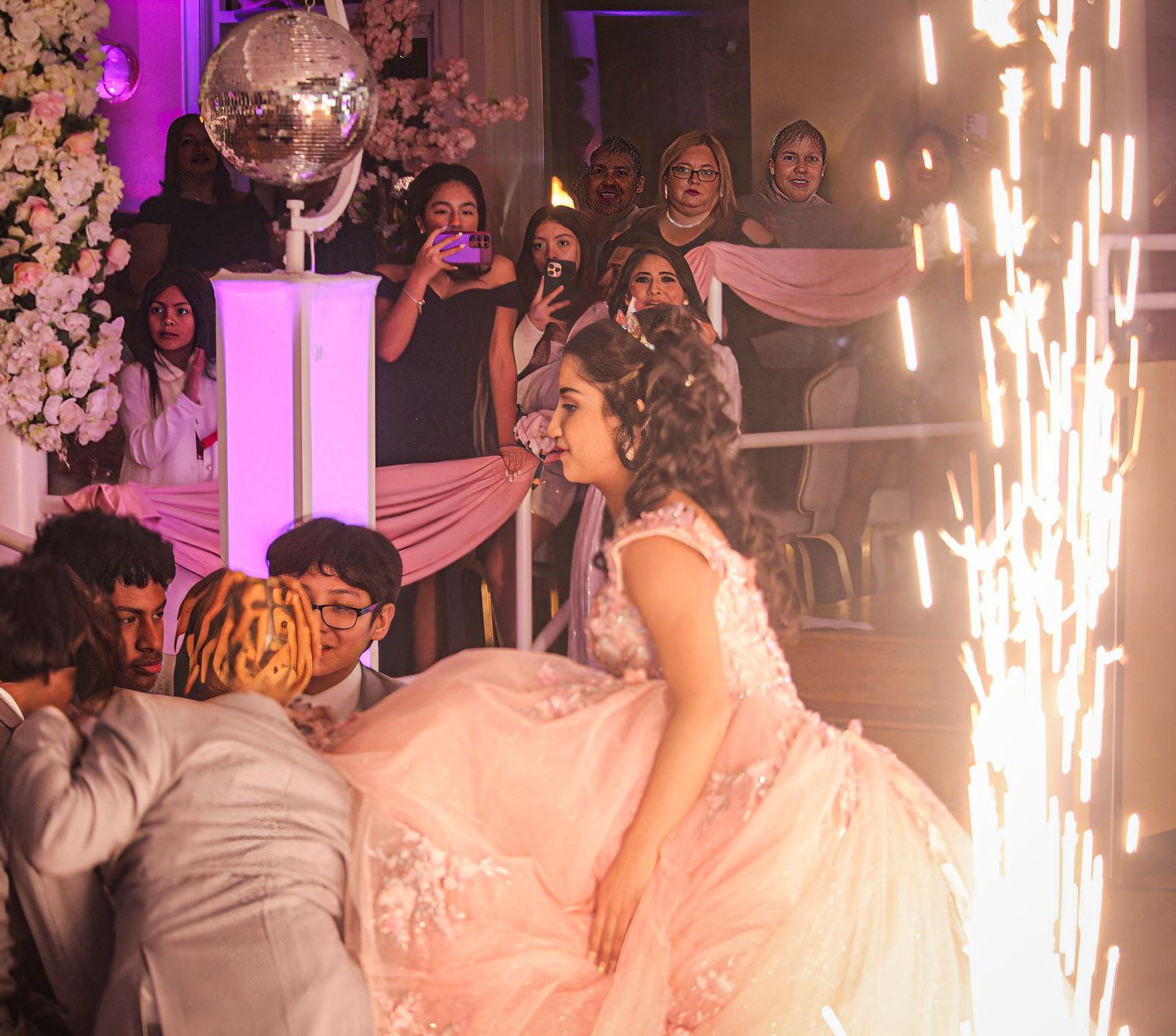 Woman in a pink gown at a celebration, with guests watching and sparkler fountains nearby