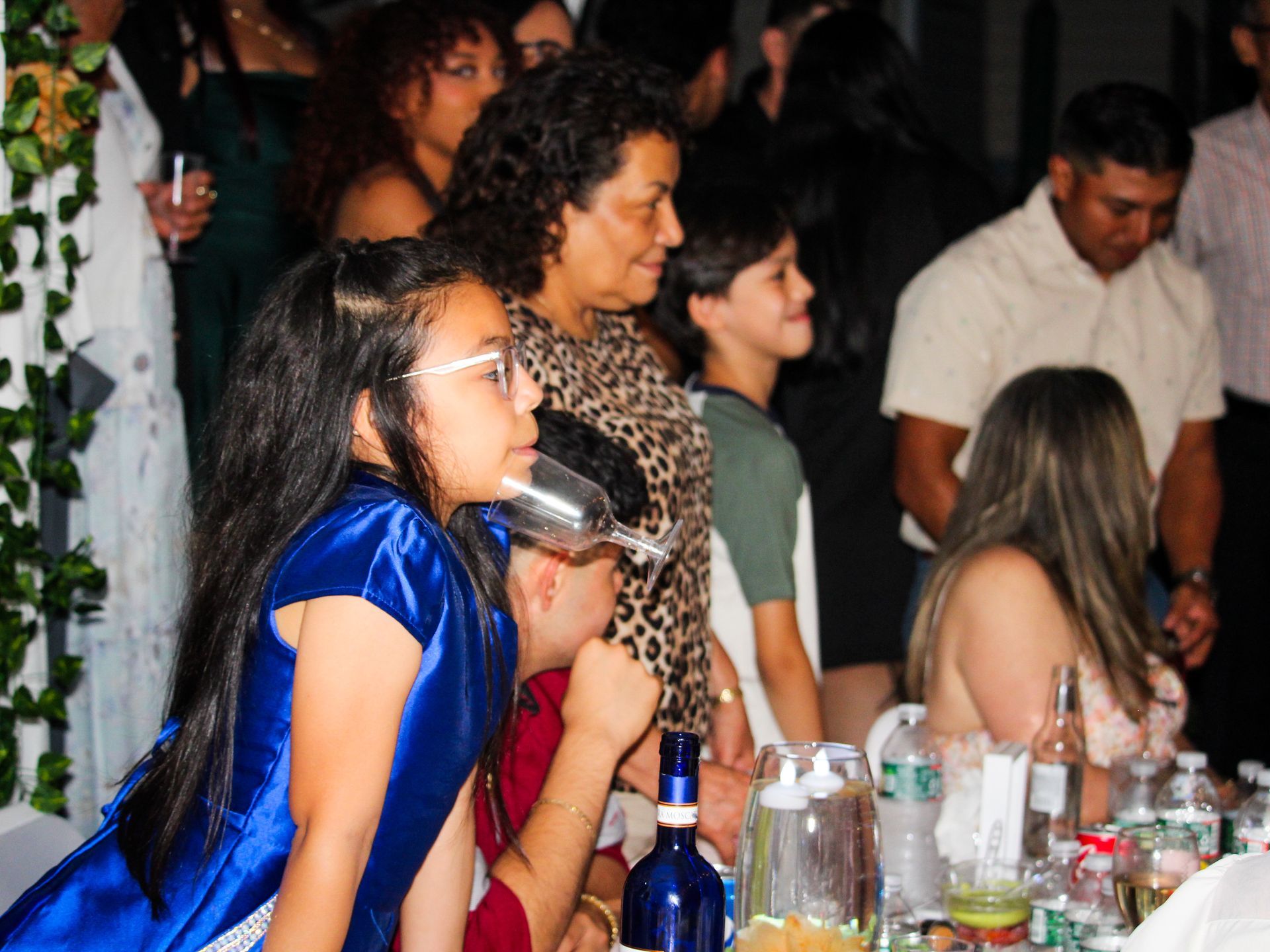 Guests gathered around a banquet table at an indoor party, with glasses, bottles, and dim warm lighting.