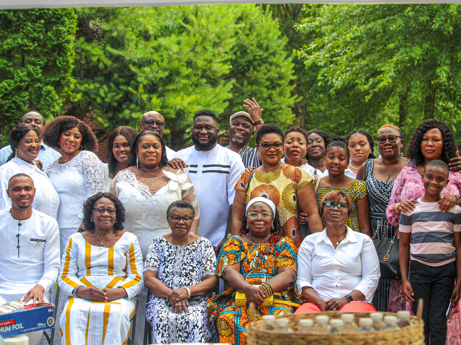Group of people posing outdoors in colorful traditional attire, smiling in front of lush greenery.